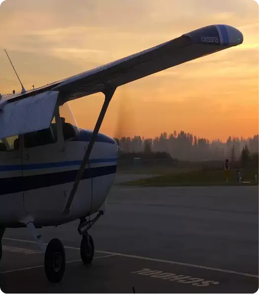 A small Cessna airplane is parked on a runway during sunset, with the orange sky and silhouettes of trees visible in the background.