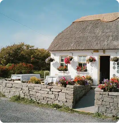 A small white cottage with a thatched roof serves as a café. Colorful flowers fill window boxes and pots on the stone patio, with outdoor seating. Trees and blue sky are in the background.