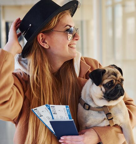 A smiling woman in a tan coat and black hat holds travel tickets and a passport in one hand and a pug dog in the other, standing indoors.