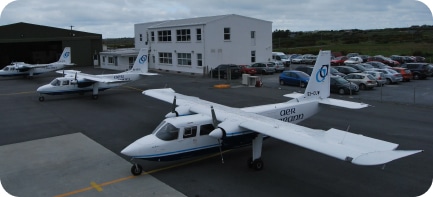 Several small twin-engine aircraft are parked on an airport tarmac near a white two-story building, with a hangar and numerous cars in the background under a cloudy sky.