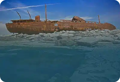 A large rusty shipwreck rests on rocky shore under a bright blue sky, with calm water in the foreground reflecting the vessel and surrounding rocks.