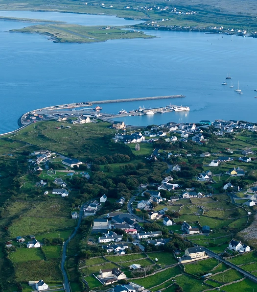 Aerial view of a coastal village with scattered houses, green fields, a harbor with boats, and a curving pier extending into blue water. Small islands are visible in the distance.