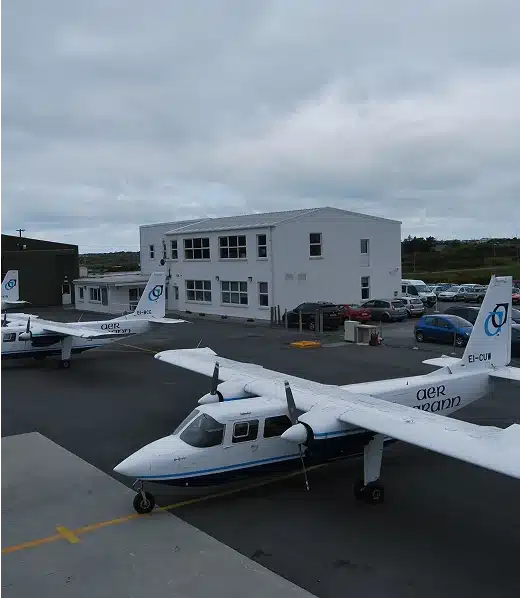 Small white airplanes are parked on an airport tarmac near a modern white building, with several cars in the parking lot and a cloudy sky overhead.