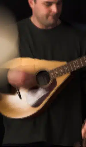 A person wearing a dark shirt plays a stringed instrument, possibly a bouzouki or mandolin. The image is slightly blurred, emphasizing the motion of the strumming hand.