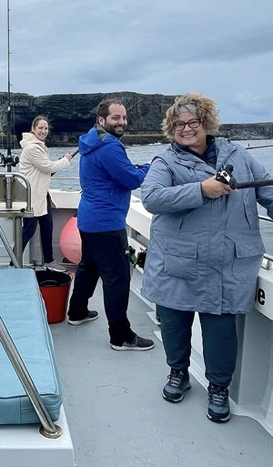 Three people in jackets stand on a boat holding fishing rods, smiling at the camera. The sky is cloudy and rocky cliffs are visible in the background across the water.