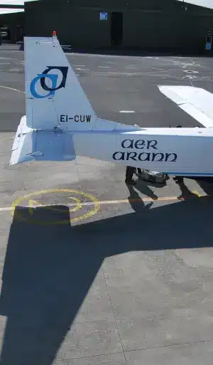Tail section of an Aer Arann airplane on an airport tarmac, with part of a hangar in the background and the aircraft's shadow visible on the ground.