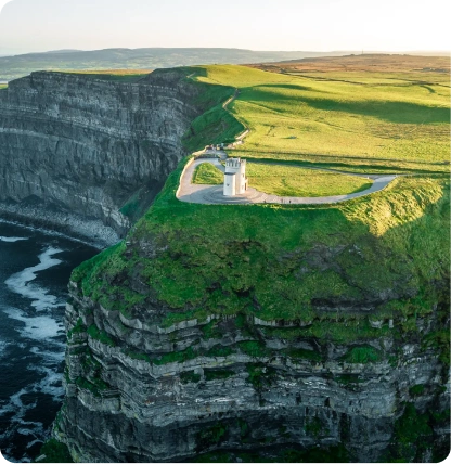 A small white tower sits atop the green, grassy Cliffs of Moher, overlooking the dark waters and dramatic rocky coastline below, under a clear sky.