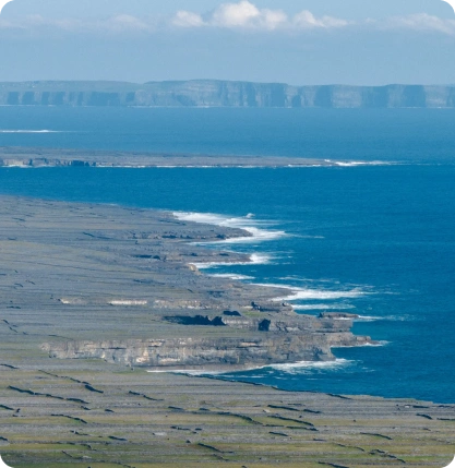 A rugged rocky coastline with waves crashing against the cliffs, stretches into the distance under a blue sky. Farther cliffs are visible across the water, partially obscured by haze.