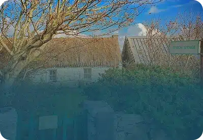 A thatched-roof cottage stands behind bare trees and bushes under a blue sky with clouds. A green sign reads “Teach Synge” near the entrance gate.