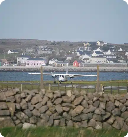 A small twin-propeller plane is taking off or landing on a rural runway, with stone walls in the foreground and a village with colorful houses in the background.