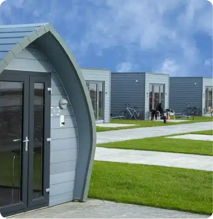 Modern, compact gray cabins with large windows are arranged in a row on green lawns, separated by concrete walkways. Several bicycles are parked outside the cabins under a partly cloudy blue sky.