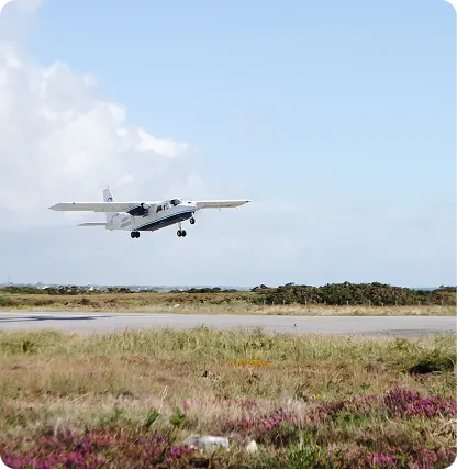 A small twin-propeller airplane takes off from a runway surrounded by grass and wildflowers under a clear blue sky.