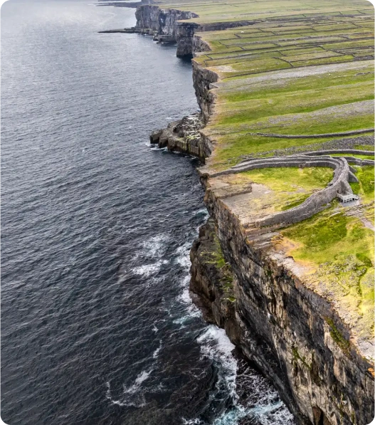 Aerial view of steep, rocky cliffs meeting the ocean, with green fields and stone walls stretching across the flat land above the cliffs. The coastline curves into the distance under a cloudy sky.