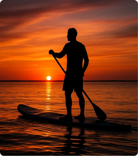 Silhouette of a person paddleboarding on calm water during a vibrant orange sunset, with the sun low on the horizon and wispy clouds in the sky.