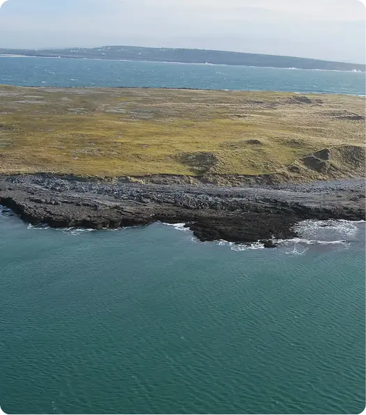 Aerial view of a rocky coastline with teal blue water, a strip of dark rocks, and grassy land extending into the distance under a partly cloudy sky.