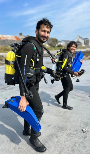 Two people in black wetsuits and scuba gear, holding blue fins, walk on a sandy beach with buildings and blue sky in the background.