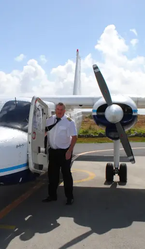 A pilot in uniform stands beside an open door of a small twin-propeller airplane on a sunny day, with one propeller and bright sky visible in the background.