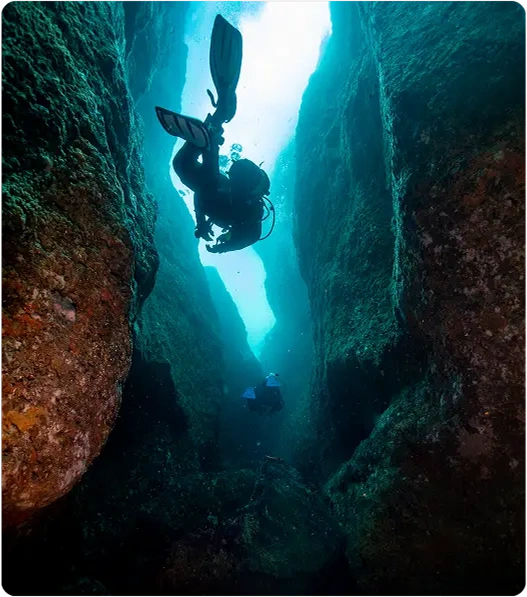 Two scuba divers swim between towering underwater rock formations in clear blue water, surrounded by rugged, algae-covered cliffs. Light filters down from above, illuminating the narrow underwater canyon.