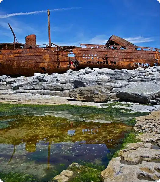 A rusted shipwreck sits on rocky shore under a bright blue sky, with green moss and a shallow tidal pool reflecting the ship in the foreground.