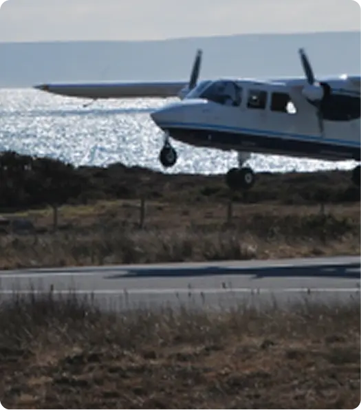 A small twin-engine plane flies low above a runway near water, with shimmering waves and distant hills visible in the background. The aircraft’s landing gear is down, preparing for landing.
