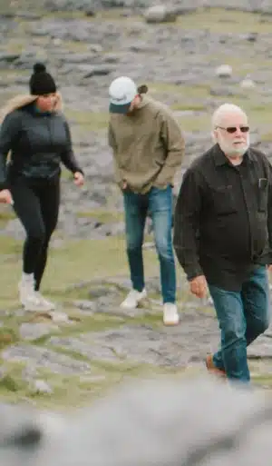 Three people walk outdoors on a rocky, grassy terrain. An older man in sunglasses and a dark jacket is in the foreground, while a woman and a man in casual clothes walk behind him.