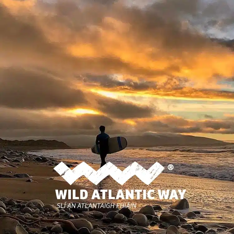 A person holding a surfboard walks on a rocky beach at sunset with clouds and waves in the background. The "Wild Atlantic Way" logo and Irish text are overlaid at the bottom.
