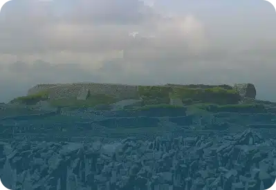 A large ancient stone fort sits on a grassy hilltop under a cloudy sky. The lower part of the image is tinted blue, partially obscuring the rocky landscape in the foreground.