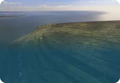 Aerial view of a coastline with patchwork fields bordered by stone walls, meeting a calm sea under a clear sky. Sunlight reflects on the water, highlighting the green and brown patterns of the landscape.