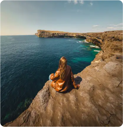 A woman with long hair, wearing a brown jacket, sits on a rocky cliff edge overlooking a calm blue sea and rugged coastline under a clear sky.
