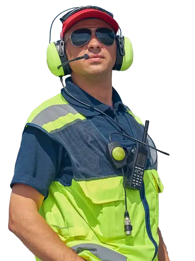 A male airport ground crew worker in a neon yellow vest, navy shirt, red cap, and headphones with a microphone, wearing sunglasses and a radio clipped to his vest.