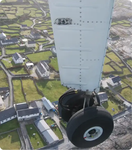 View from an airplane showing a close-up of the aircraft’s landing gear and wing, with green fields, stone walls, and houses visible below.