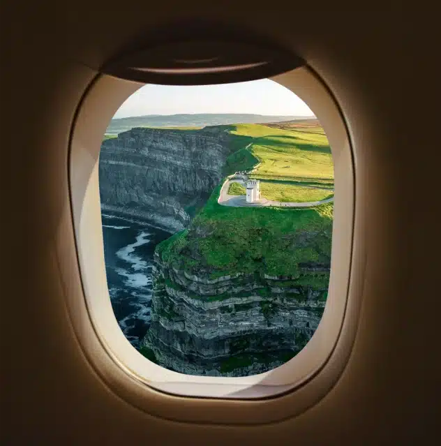 View through an airplane window of dramatic, steep green cliffs overlooking the ocean, with a small lighthouse perched near the edge and blue sky above.