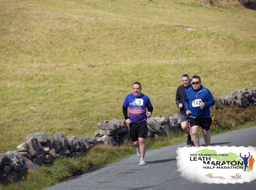 Three men run along a rural road bordered by a stone wall during the Leath Marathon Half Marathon, with grass fields in the background. Event logo and text appear in the bottom right corner.