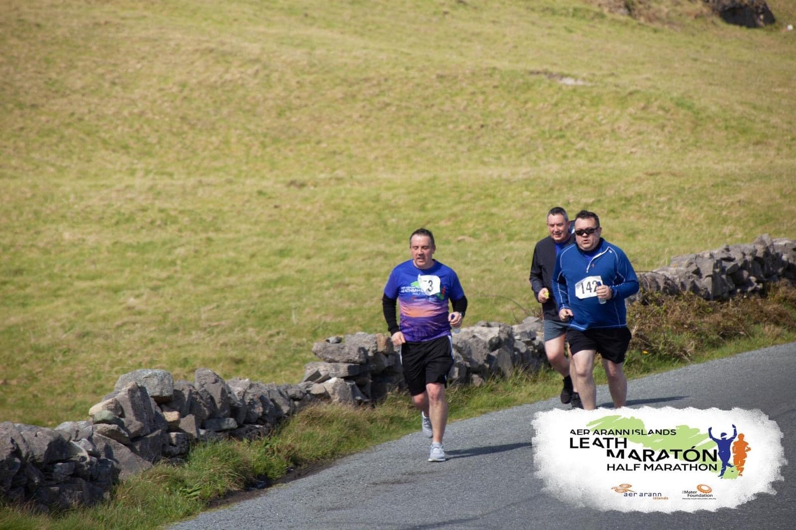 Three men run along a rural road bordered by a stone wall during the Leath Marathon Half Marathon, with grass fields in the background. Event logo and text appear in the bottom right corner.
